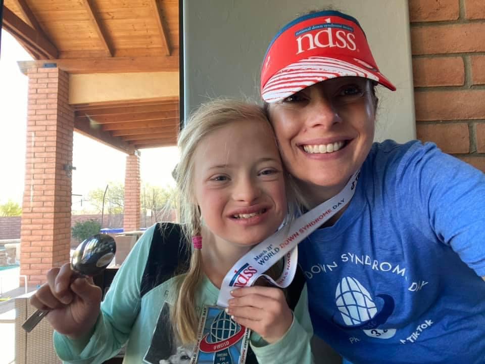 Girl with Down syndrome and her mother smile at camera wearing racing for 3.21 hat, shirt, and medal
