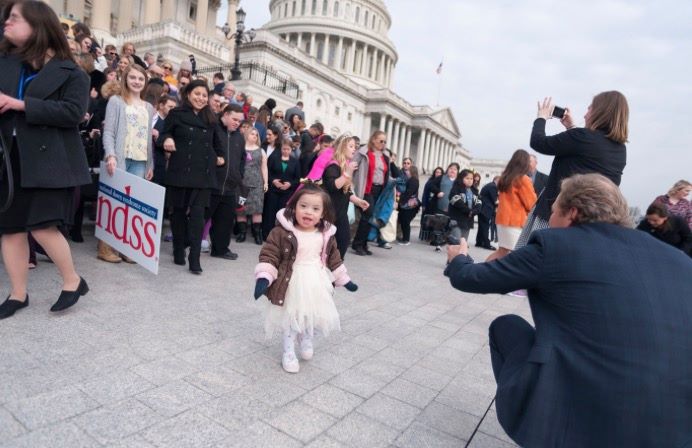 Image of little girl with Down syndrome running to the camera in front of a crowd of the Down syndrome community in front of the capitol