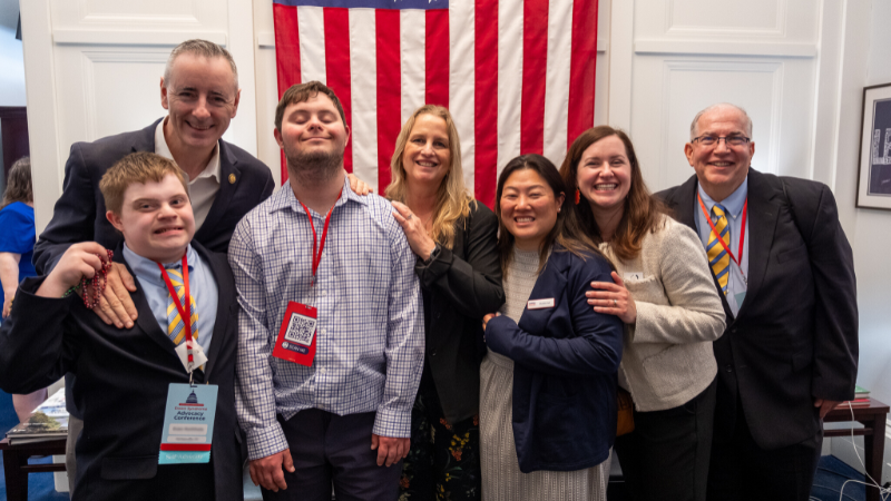 A group of advocates with and without Down syndrome meet in the Capitol Building in front of an American flag.