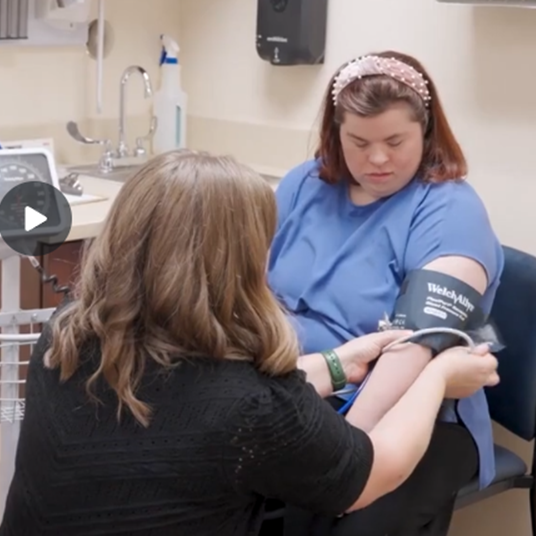 A young, white woman with Down syndrome is seated in a medical exam room having her blood pressure taken by a white woman with ligh brown hair. Her back is to the camera. The woman with Down syndrom has light brown hair and is wearing a pink headband and blue shirt with dark slacks. 