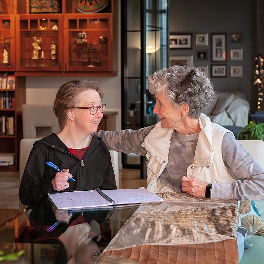 An older white woman with Down syndrome who has short brown hair and is wearing glasses and a dark shirt is sitting in front of some papers at a table and holding a pen. She is seated next to an older white woman with short gray hair with her arm around the back of the chair of the woman with Down syndrome. They are both smiling and looking at each other. 