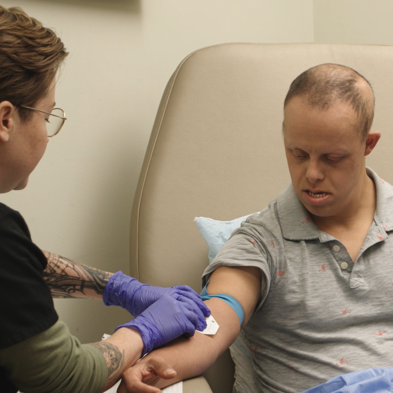 A white man with Down syndrome is seated in an exam chair wearing a gray polo shirt. He is having his blood drawn by a white woman with short auburn hair and glasses, wearing scrubs and purple exam gloves. 