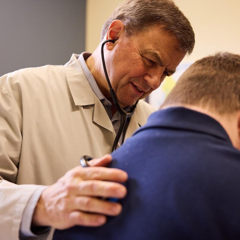 A middle-aged man in a white coat with a kindly face and a stethoscope in his ears has his hand on the back of a young man wearing a blue shirt. 