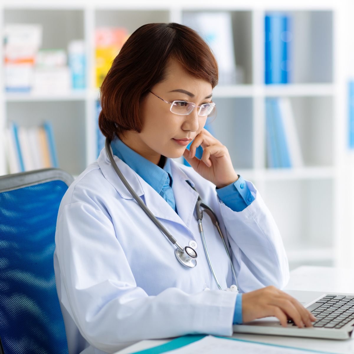 An Asian woman with short brown hair is wearing glasses, a white lab coat, and a stethoscope around her neck. She is at a desk with a keyboard and is reading a screen.