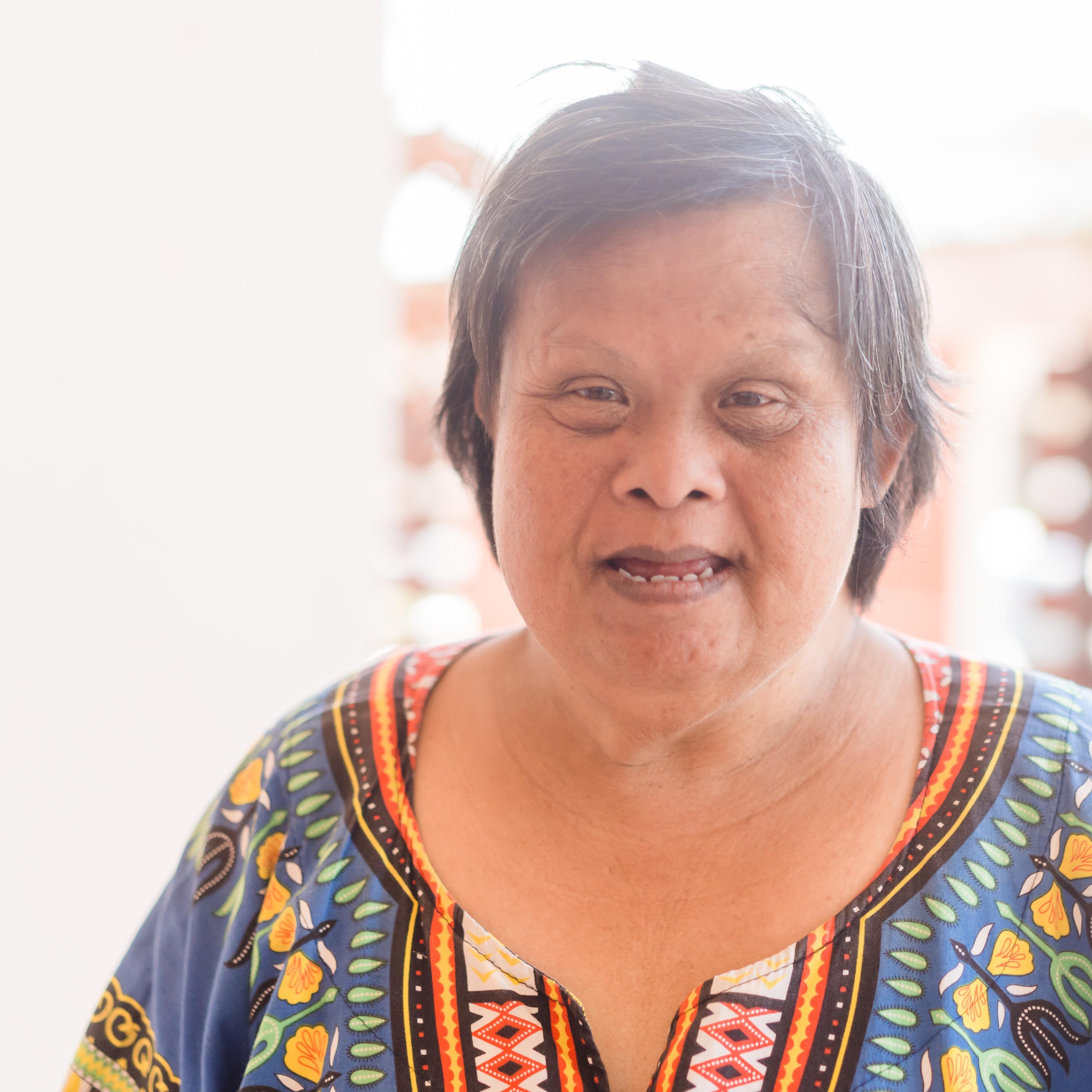An older woman with Down syndrome is wearing a colorful shirt and has short, graying hair. She is standing in front of a window, and smiling directly at the camera.