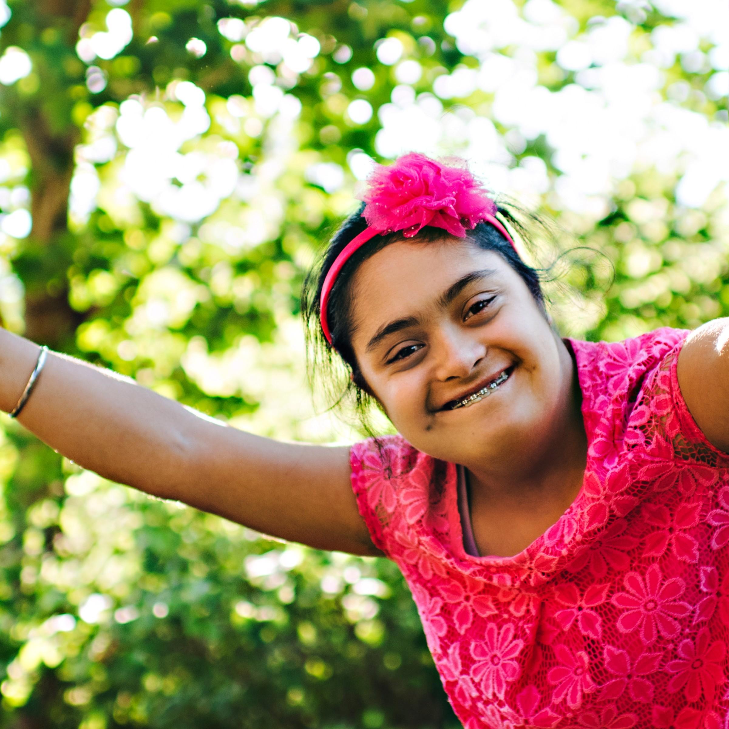 A young woman with Down syndrome is outside with trees behind her. She is wearing a hot pink shirt and bow in her hair. She has braces on her teeth, and she is smiling directly at the camera.