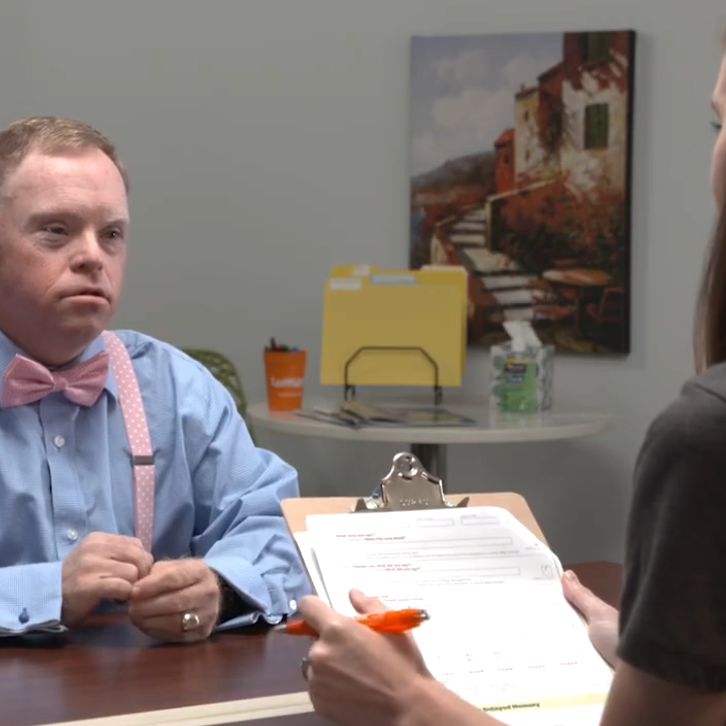 An older man with red hair, who has Down syndrome is wearing a blue shirt and a pink bow tie. He is sitting at a table, opposite a woman with brown hair who is holding a clipboard. 