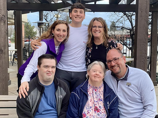 A group of people are smiling with their arms around each other in front of a covered outdoor area. In the front of the group from left to right there is a young, dark-haired white man with Down syndrome seated next to an older white haired, white woman with Down syndrome and a middle aged white man with a bald head and gotee. Behind them from left to right are a young white woman with long sandy hair, a young white man with dark hair and a middle-aged white woman with light brown hair. 
