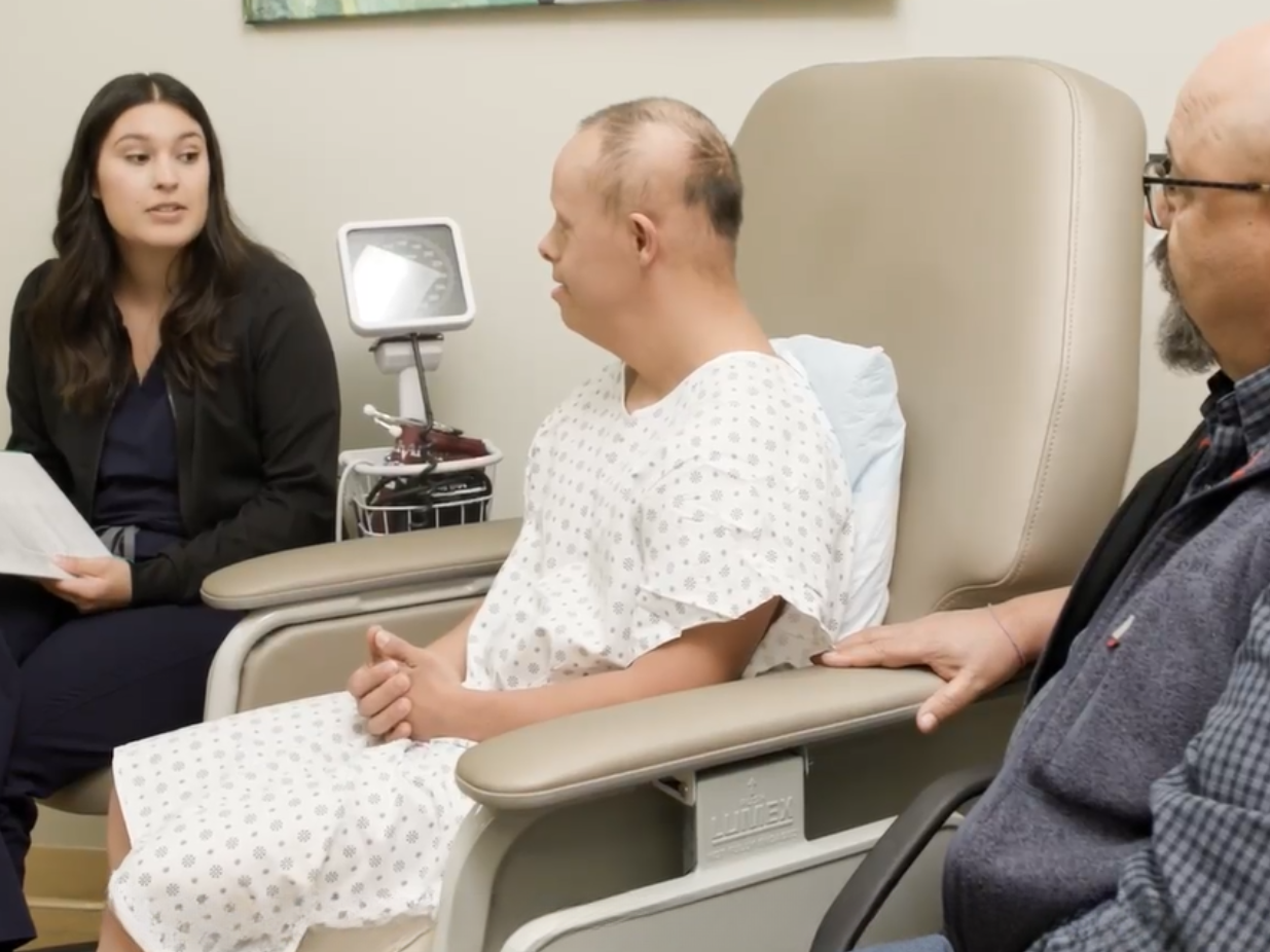 A white man with Down syndrome is seated in an exam chair wearing a hospital gown. Next to him on one side is a young woman with dark hair who is seated and is speaking with papers in her hand and on the other side is a bald man with glasses and a gotee. The men appear to be listening to what the woman is explaining to them.  