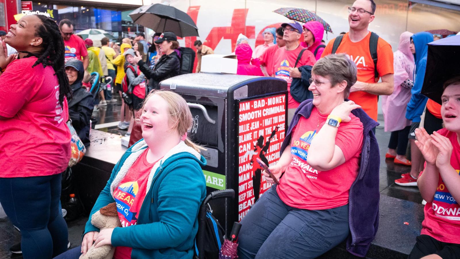 Blonde woman with Down syndrome, using a wheelchair, smiles at the Times Square billboard