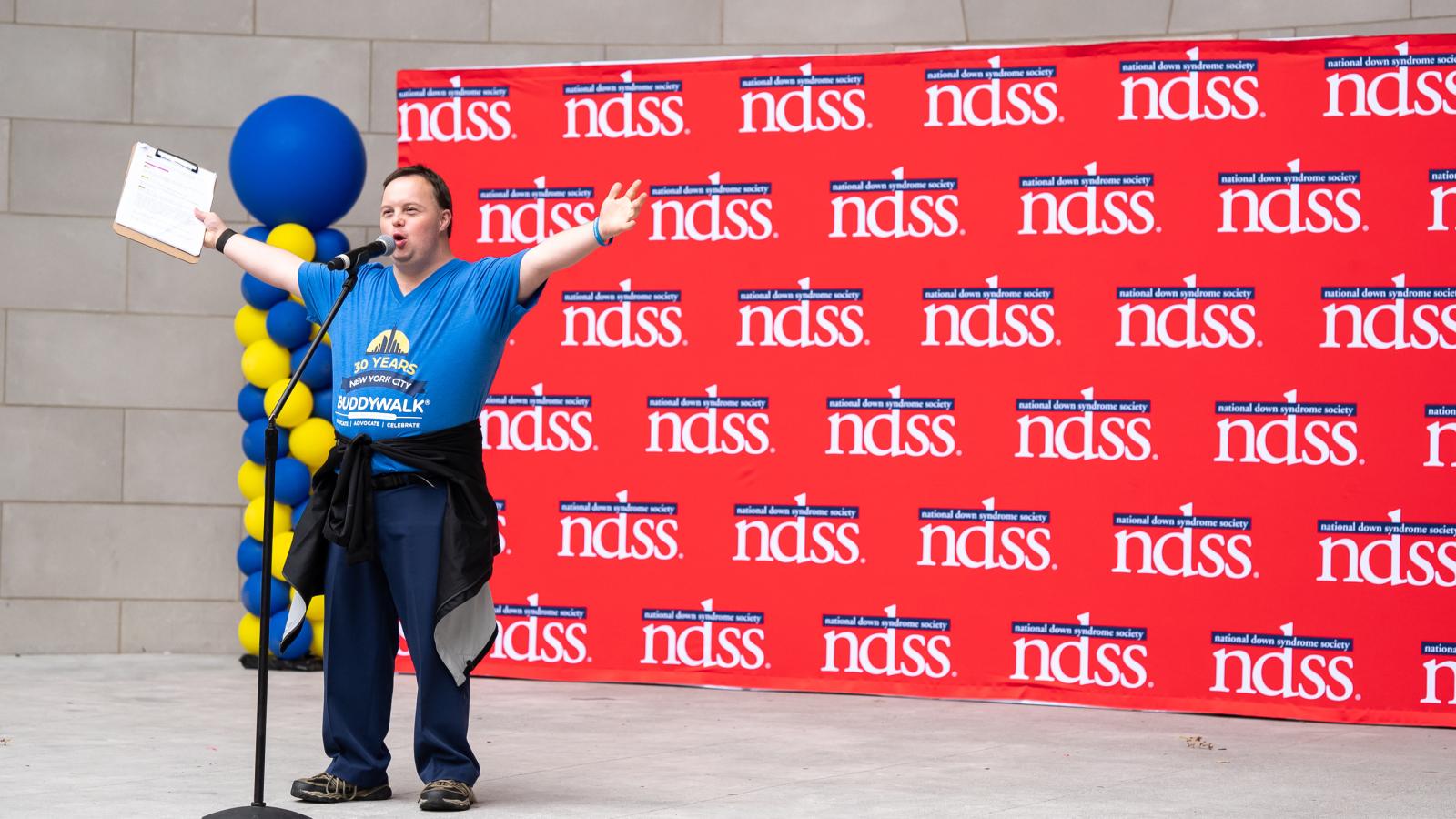 David, a man with Down syndrome and NDSS staff member, welcomes a crowd from a stage in front of an NDSS banner