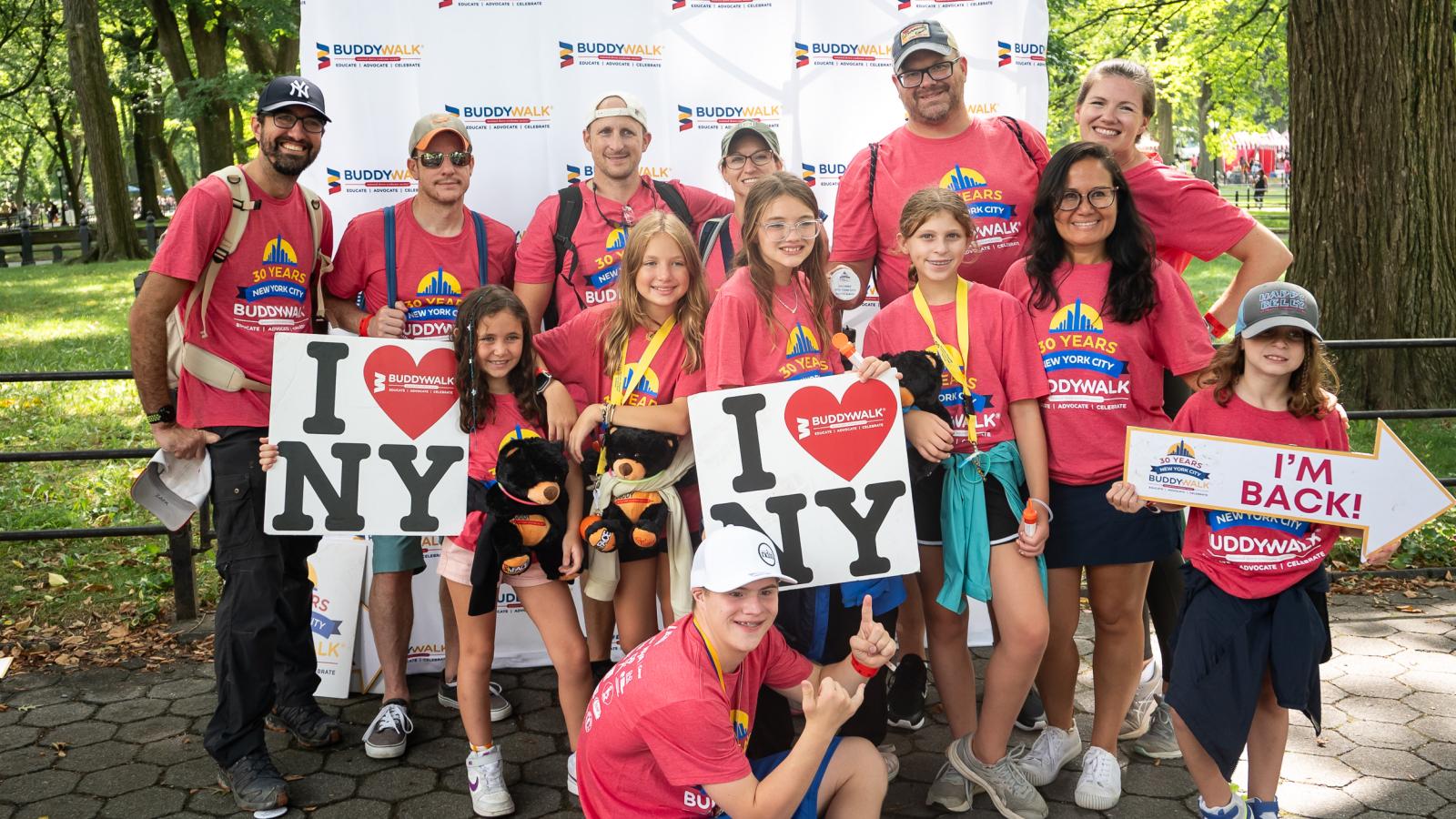 A group, wearing red Buddy Walk shirts, holds up I heart NYC signs