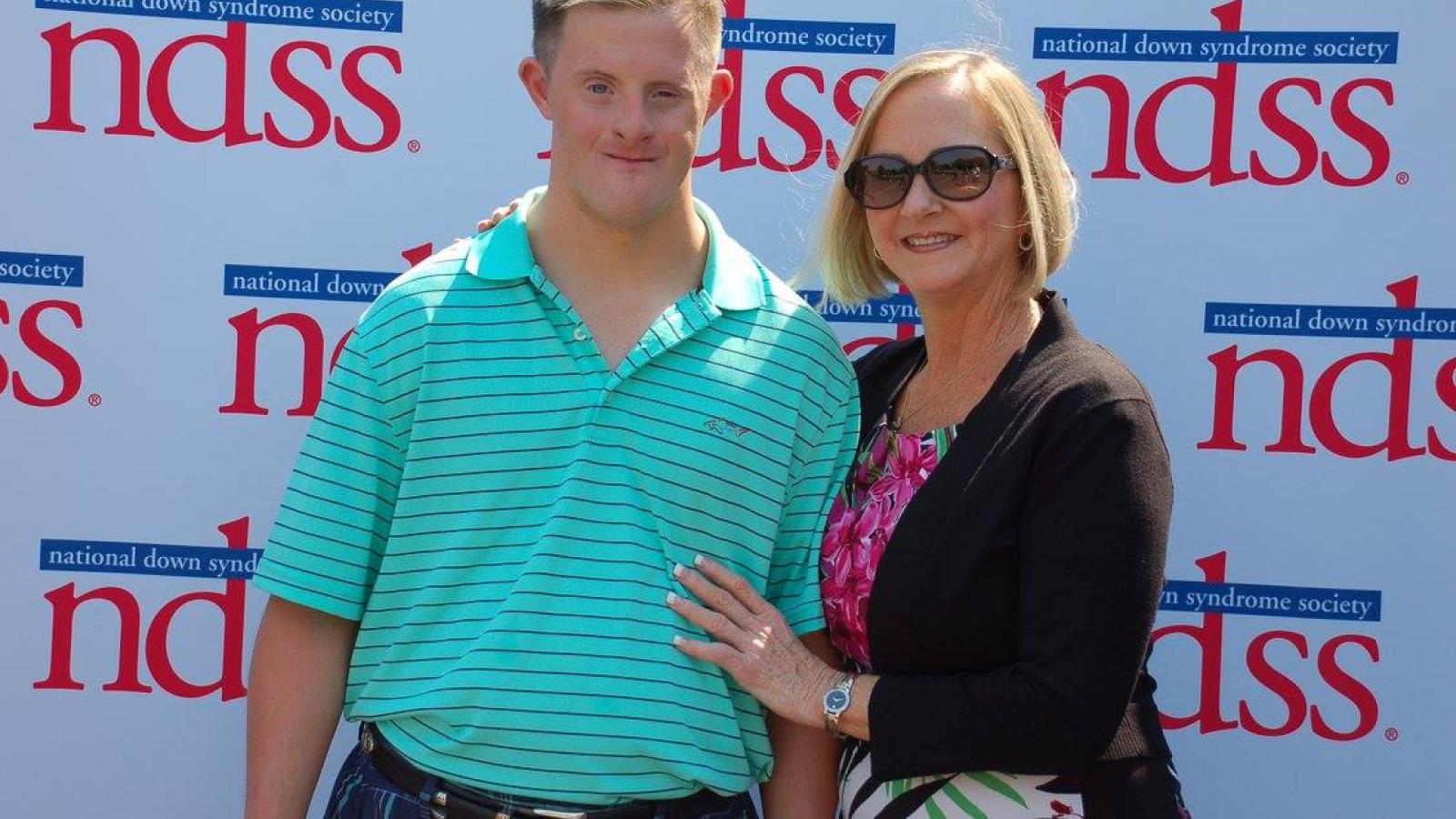 A man with Down syndrome and a woman smile in front of an NDSS banner