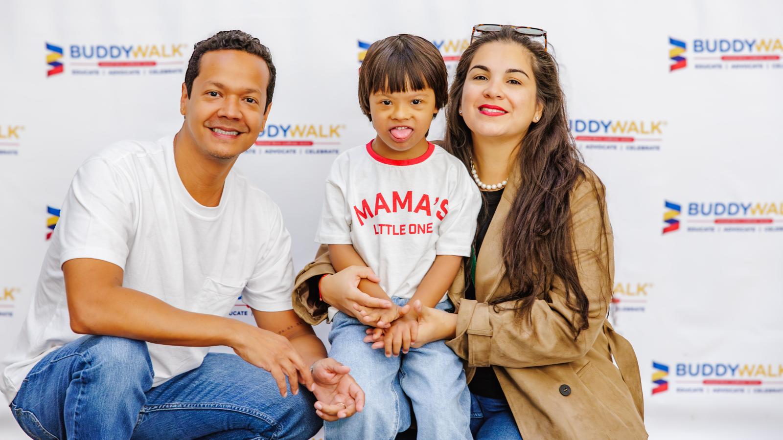 A family with a young boy with Down syndrome poses in front of a Buddy Walk banner