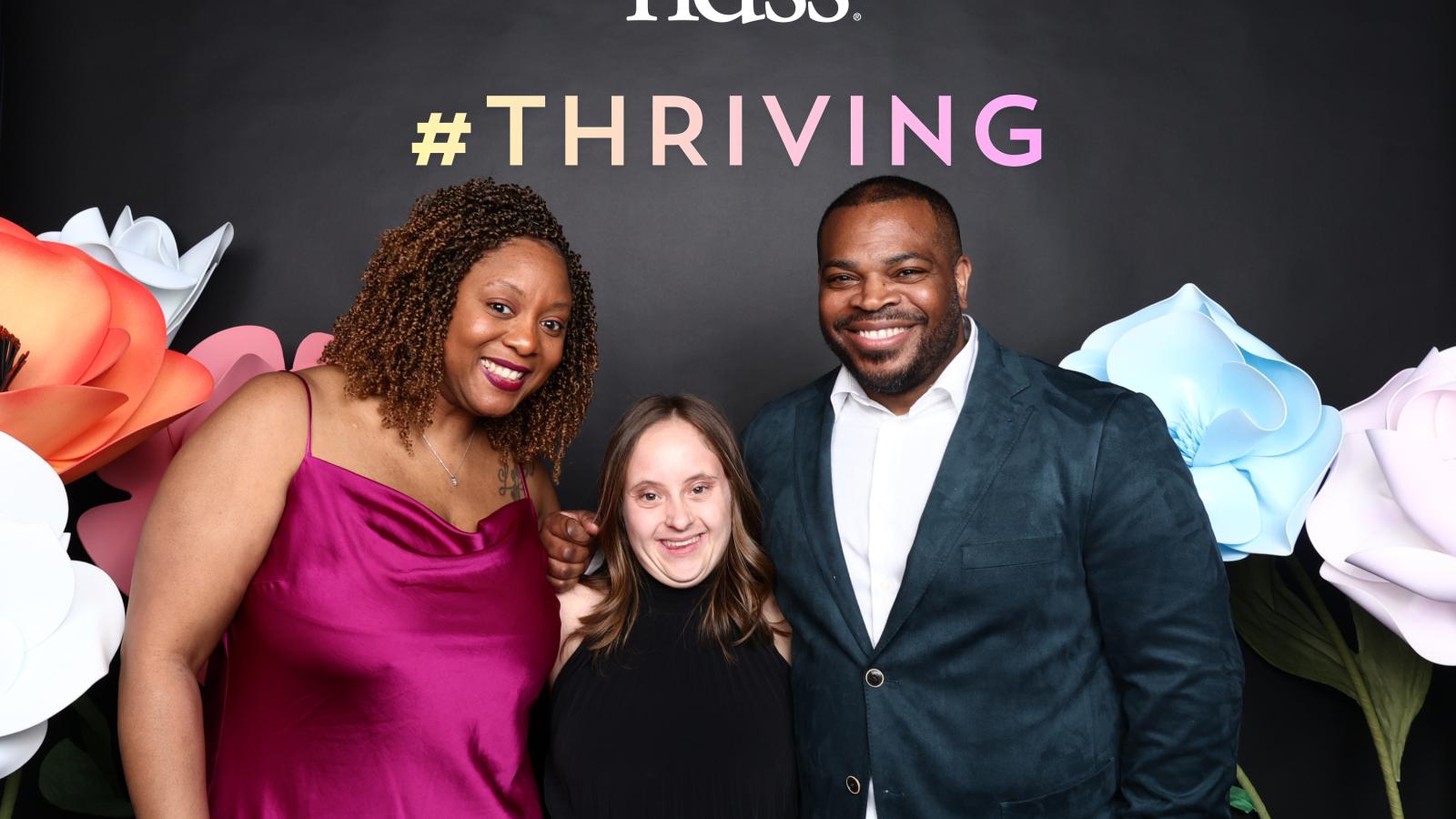 Three people smile in front of a black back drop with flowers, the NDSS logo, and "#THRIVING"