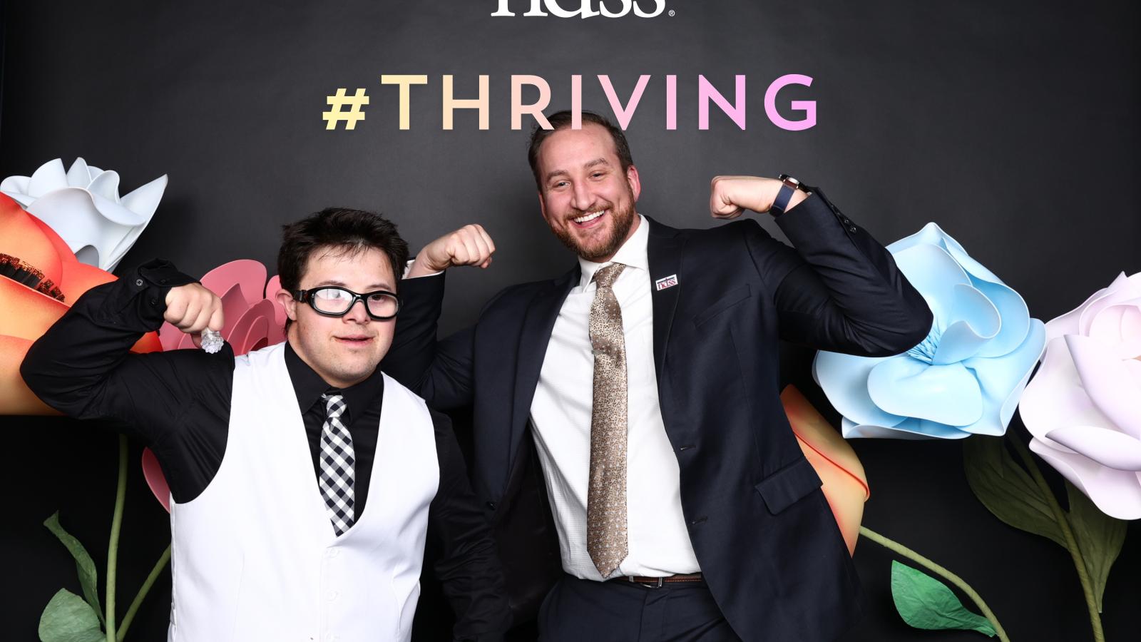 A young man wearing a vest and tie poses with another man wearing a suit. They pose to show that they are strong in front of a black back drop with flowers, the NDSS logo, and "#THRIVING"