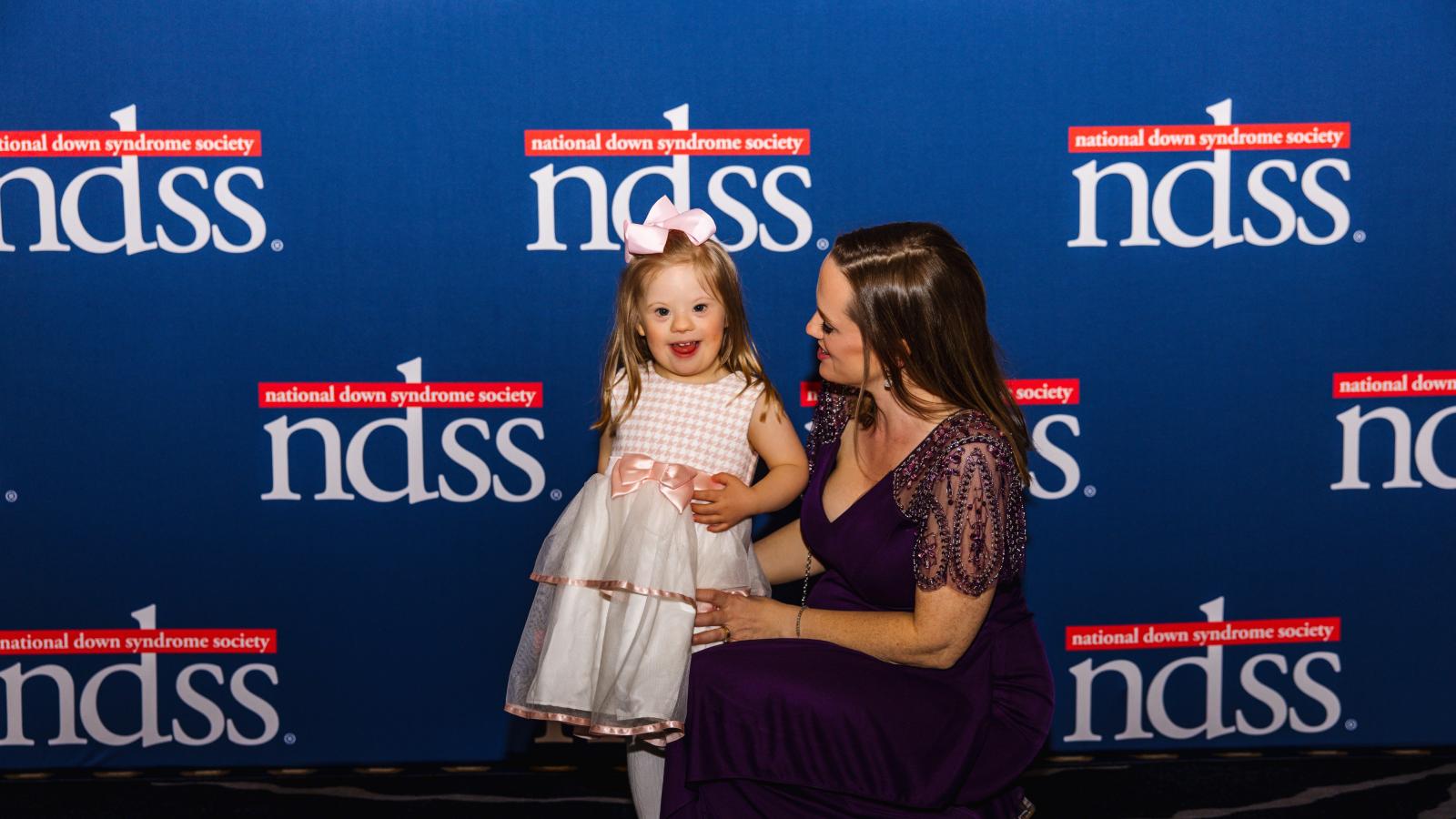 A woman poses with a young girl with Down syndrome in front of a blue back drop with NDSS logos