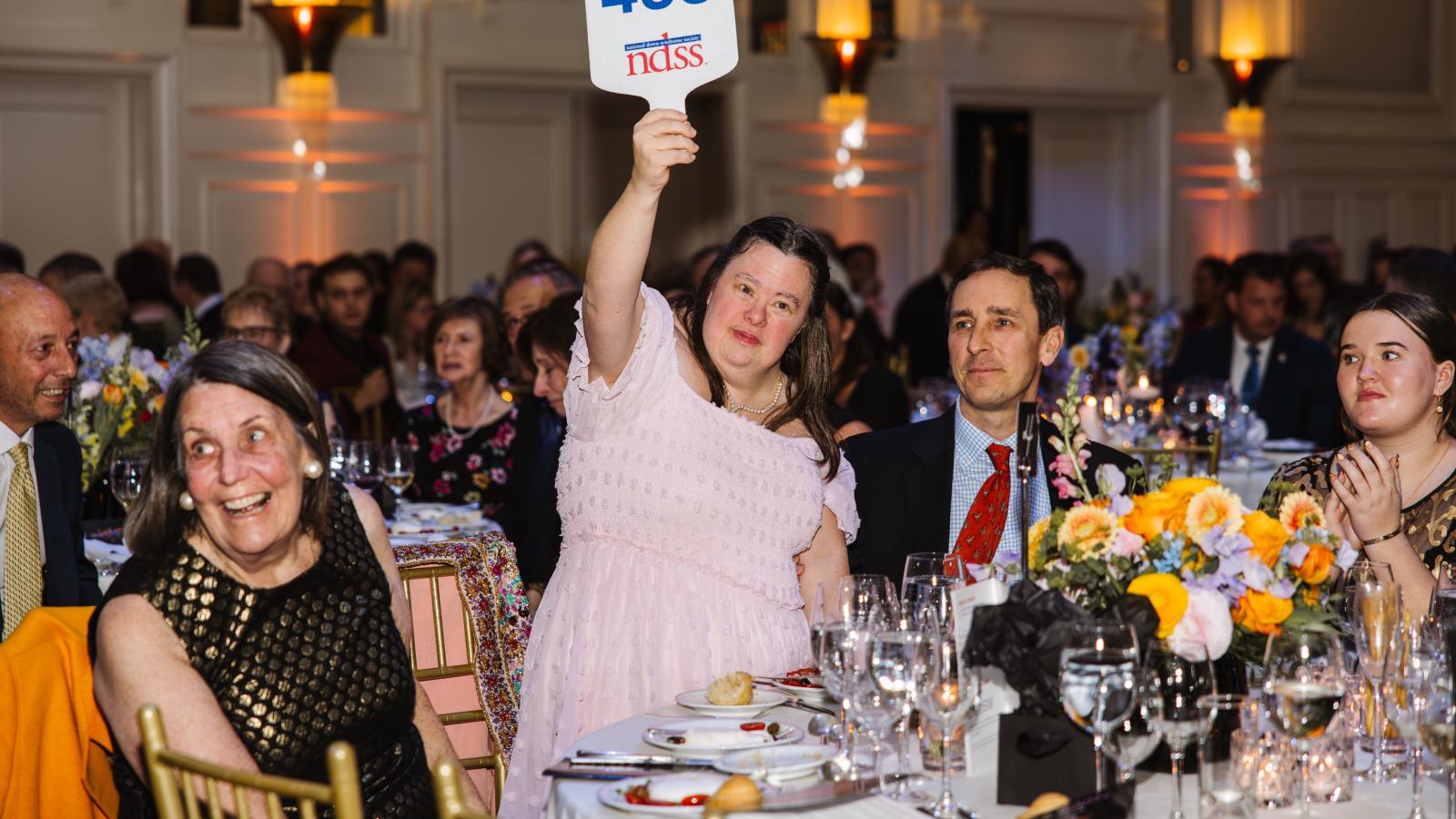 A woman with Down syndrome, wearing a pink gown, holds up sign at a paddle raise
