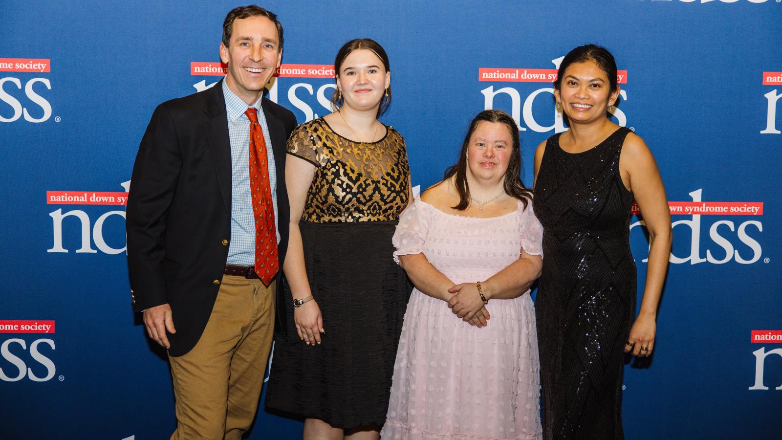 A group of people, including a woman with Down syndrome, pose in front of a blue banner with NDSS logos.