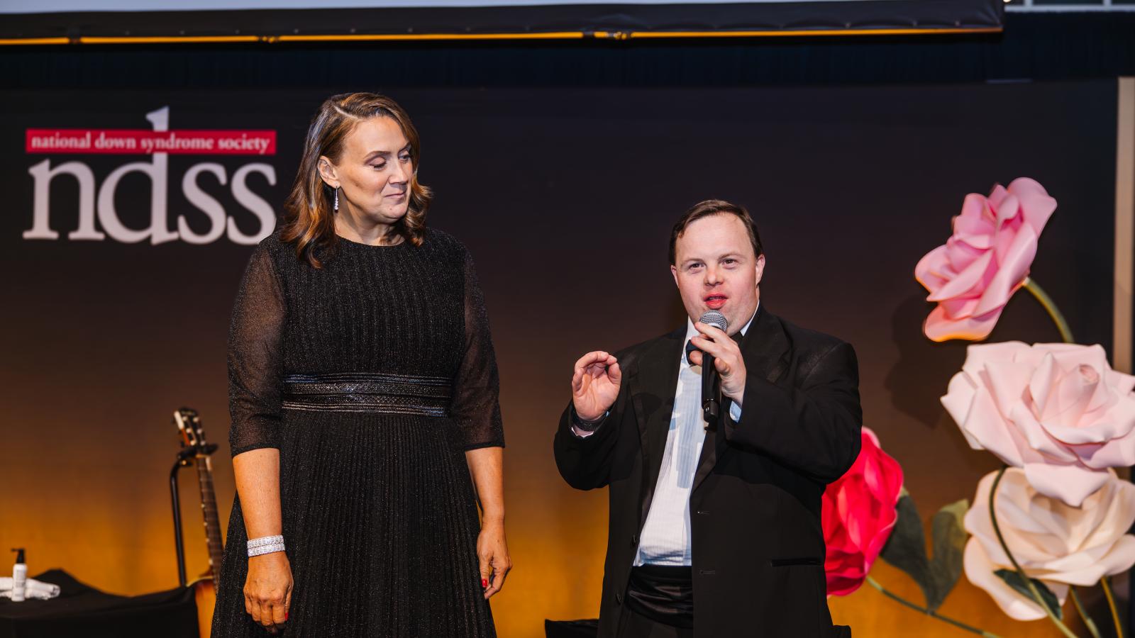 NDSS CEO Kandi Pickard stands on stage while David DeSanctis, a man with Down syndrome and NDSS Development Outreach Coordinator, speaks into a microphone