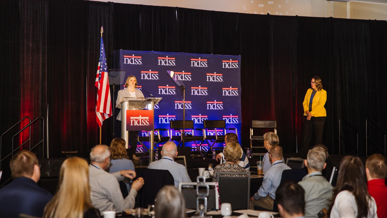 A woman with Down syndrome stands at a podium bearing the NDSS logo on a stage in front of a seated audience at the Down Syndrome Advocacy Conference. An American flag stands to the left of the podium, and a second speaker in a yellow blazer stands on the stage to the right. An NDSS step-and-repeat backdrop is in the background.