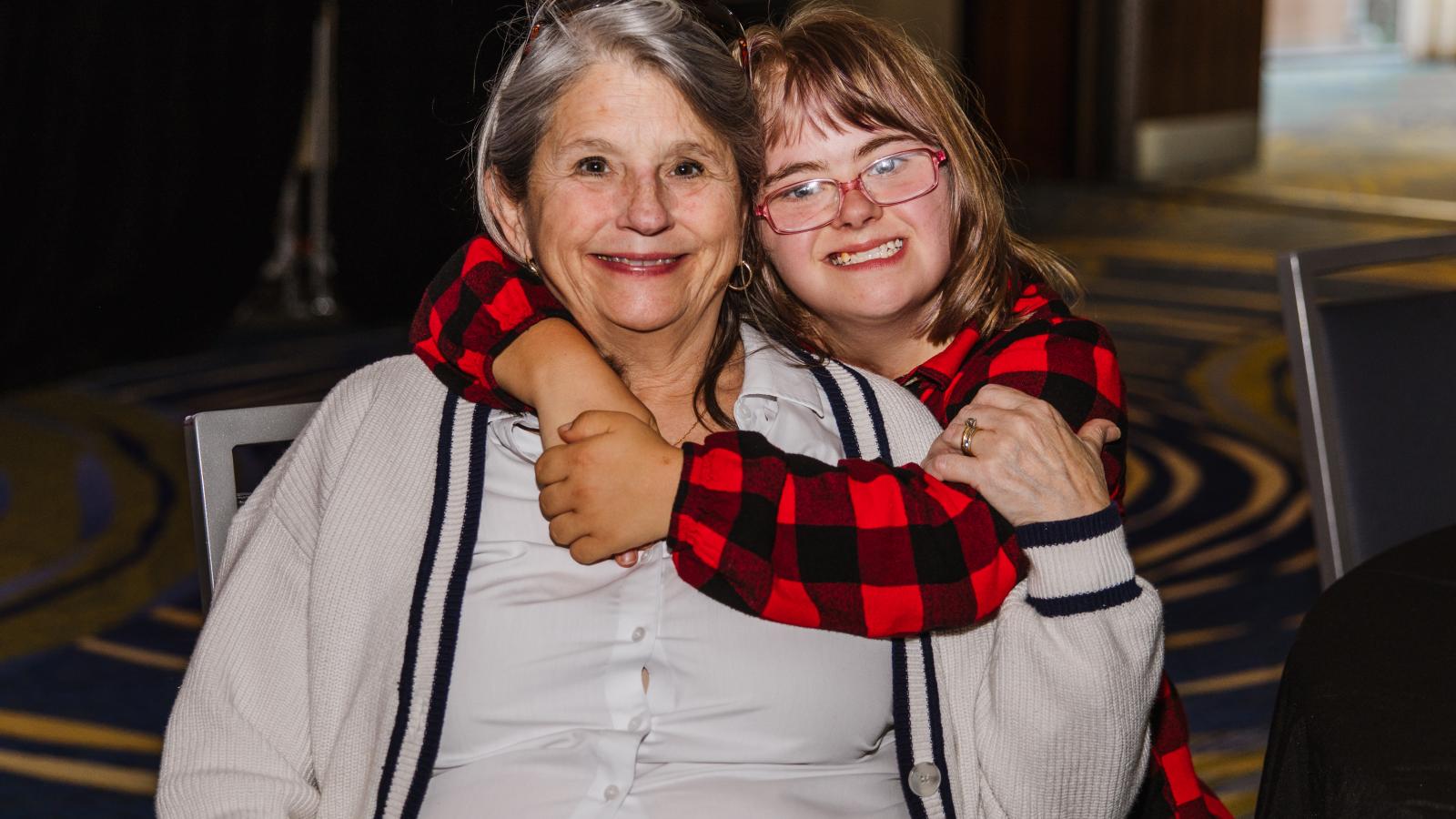  Two women sit closely together, smiling at the camera. The younger woman, who has Down syndrome, is wearing a red plaid jacket and glasses, and has her arms wrapped warmly around the older woman, who is wearing a white collared shirt and suspenders...