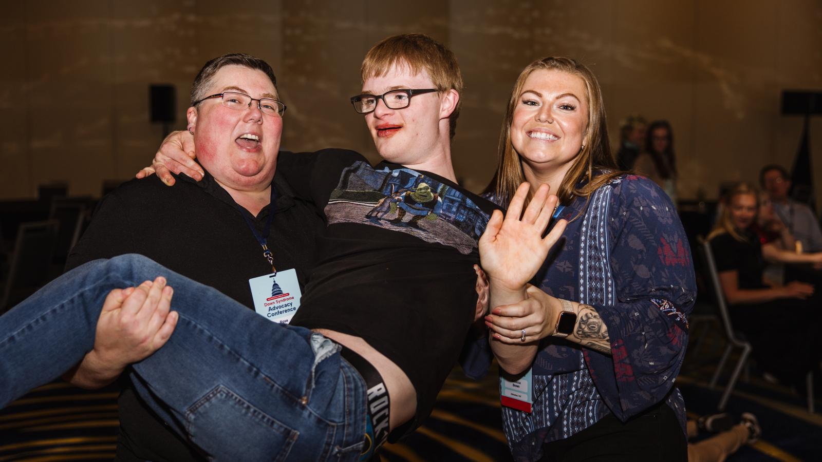 Three conference attendees — two adults and a young man with Down syndrome — share a fun, playful moment. One adult is holding the young man in their arms while all three laugh and wave at the camera. Conference lanyards are visible.