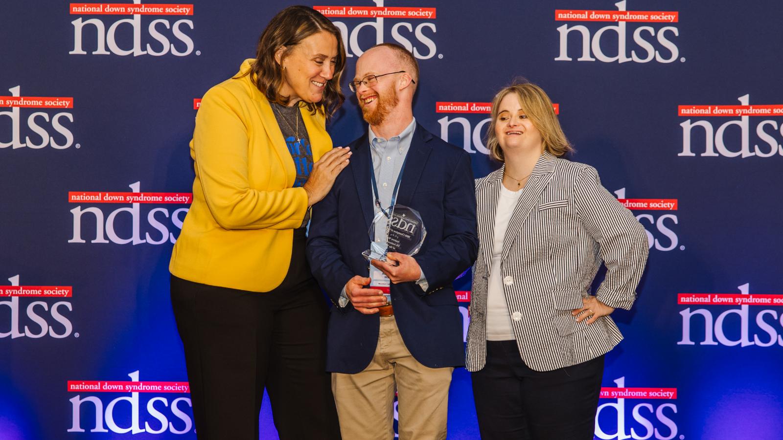 Three individuals stand in front of an NDSS step-and-repeat backdrop. A man with Down syndrome, wearing a blue blazer, holds a glass NDSS award and laughs as the tall woman to his left, wearing a yellow jacket, smiles at him. A woman with Down syndrome in a striped blazer stands to his right.