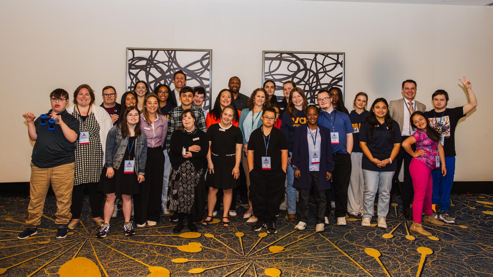 A large group of approximately 25 young adults and teens poses together in a conference room. Many are wearing conference lanyards and business-casual attire. Several individuals with Down syndrome are among the group; some are making playful gestures and peace signs.