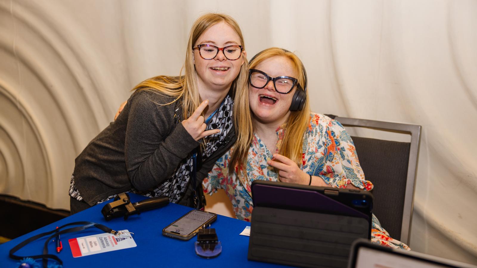 Two young women with Down syndrome and blonde hair lean together and smile broadly at a registration table. One is wearing headphones and a floral top, and the other is making a "rock on" hand sign. A tablet, phone, and NDSS badge are visible on the blue-draped table in front of them.