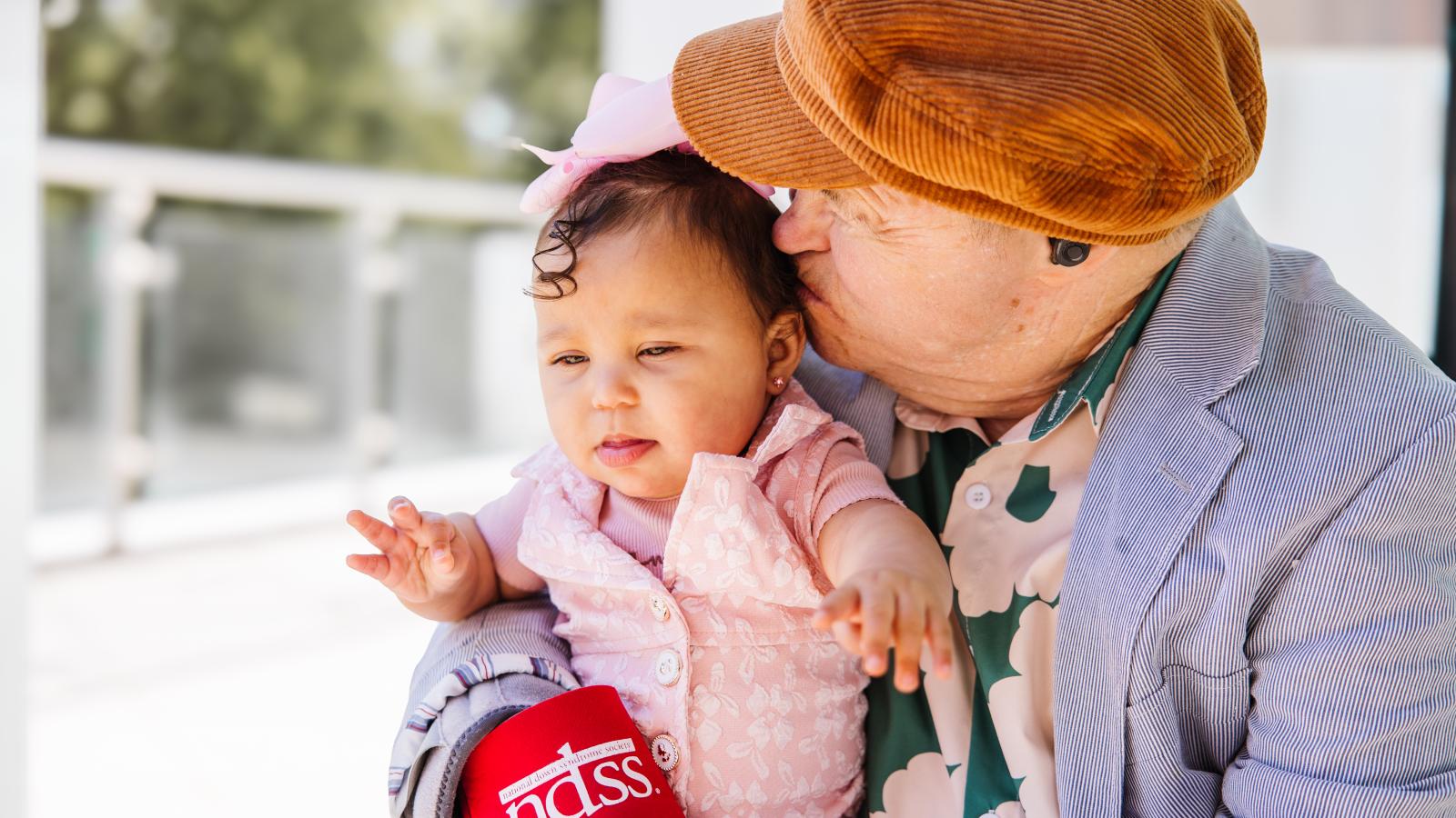 An older individual with Down syndrome, wearing a brown newsboy cap, a green shirt with pink flowers, and a suit jacket, gently kisses a baby dressed in pink. The older individual is holding a red NDSS branded cup. The photo is taken outdoors on a patio.