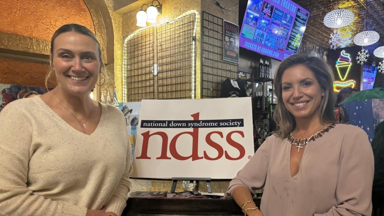 Two women stand smiling on either side of am NDSS (National Down Syndrome Society) sign inside a theater lobby decorated with snowflakes.