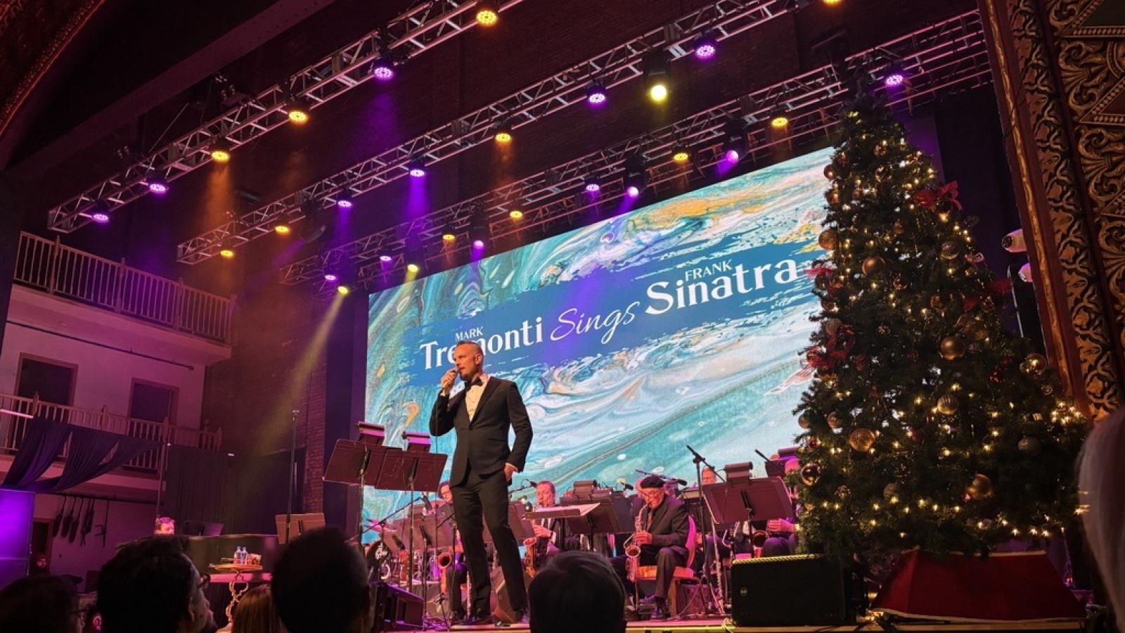 A man in a suit sings into a microphone on a stage decorated with a Christmas tree, with a screen behind him reading "Mark Tremonti Sings Frank Sinatra" and a band on the stage.