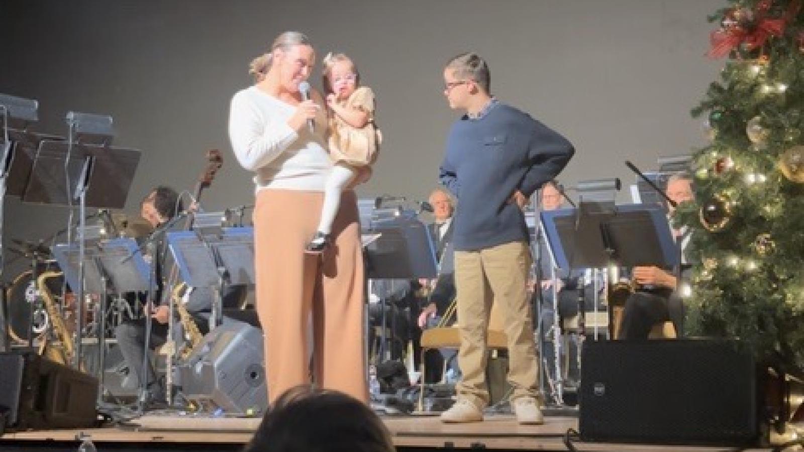 A woman holds a young girl with Down syndrome while standing on a stage next to a boy with Down syndrome, with a band and Christmas tree in the background and an audience seated below.
