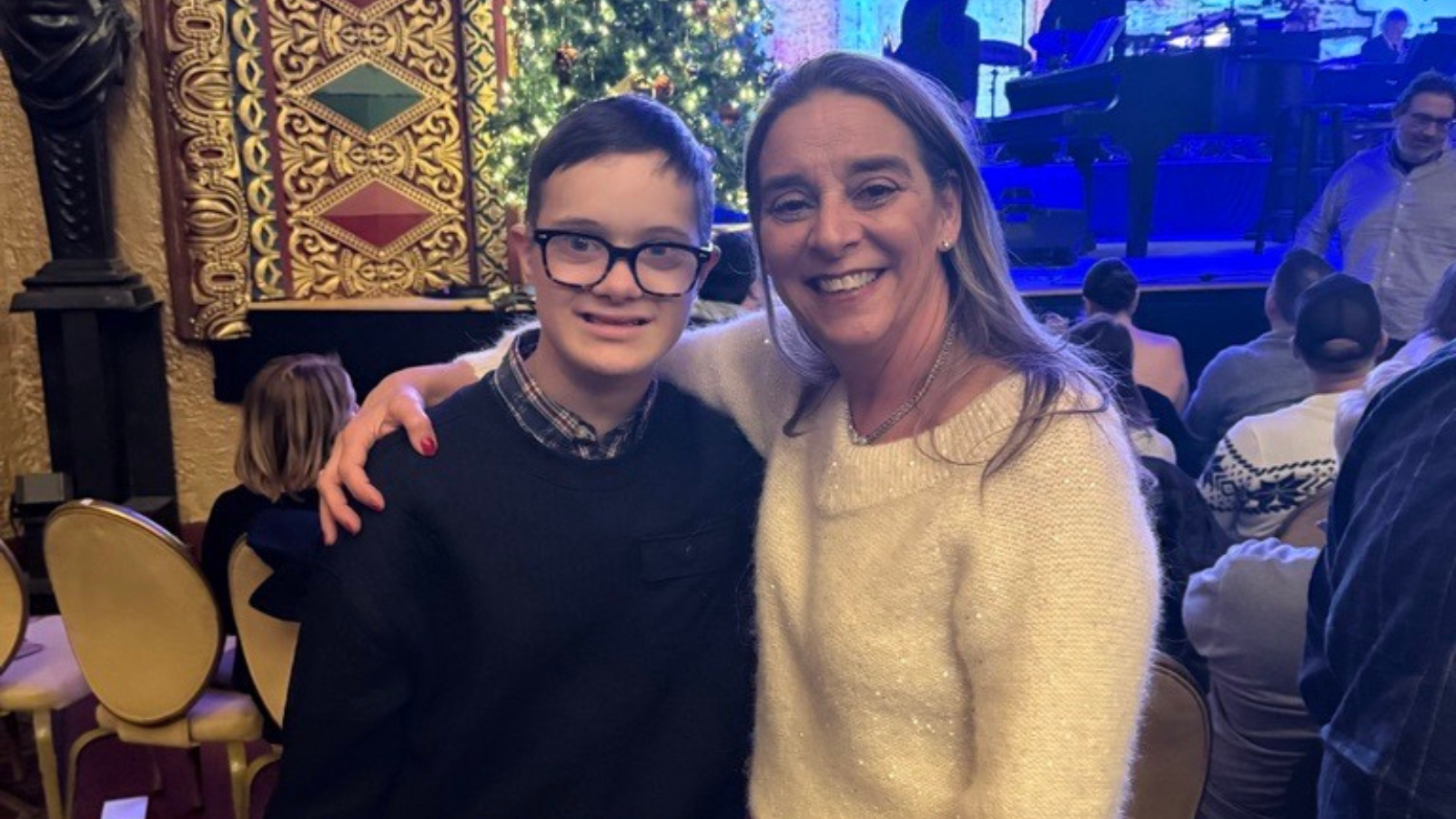 A boy with Down syndrome, wearing glasses, smiles with his arm around a woman in a cream sweater, standing near a Christmas tree in a decorated theater.