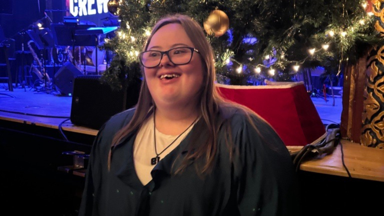 A woman with Down syndrome, wearing glasses, smiles in front of a lit Christmas tree on a stage.