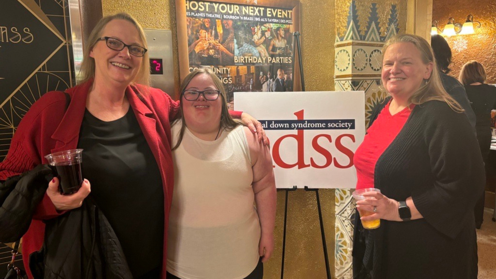 Two women stand on either side of a woman with Down syndrome, posing next to a National Down Syndrome Society sign in a theater lobby.
