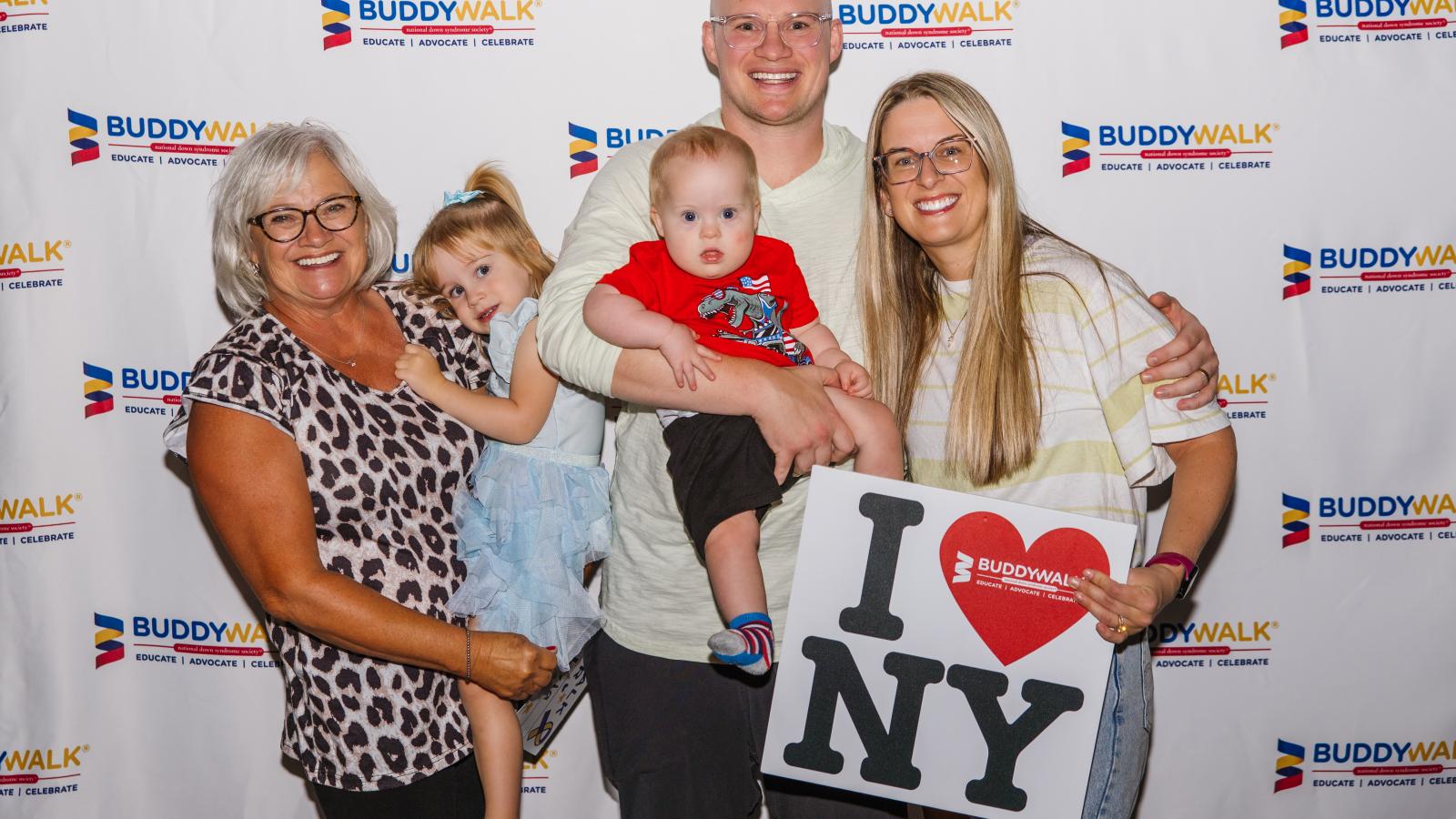 A woman with gray hair and glasses in a leopard-print top stands next to a girl in a light blue ruffled dress, a man in glasses and a cream sweater holding a baby boy with Down syndrome wearing a red dinosaur T-shirt, and a woman in glasses with a striped top holding a sign that reads "I [heart] NY" with a Buddy Walk logo inside the heart. They pose in front of a white backdrop repeating the Buddy Walk logo (a blue, gold, and red ribbon icon) with the text "Buddy Walk, National Down Syndrome Society"
