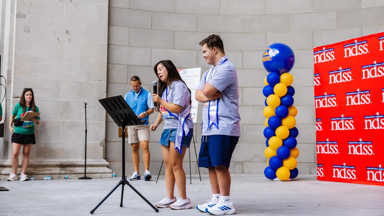 A young woman with Down syndrome, wearing a gray T-shirt, denim skirt, and a blue-and-white sash, speaks into a microphone on a stand. Beside her, a young man in a matching gray T-shirt and sash stands with his arms crossed, smiling. They stand in front of a tall stone wall near a red backdrop repeating the NDSS logo and a column of blue and gold balloons.