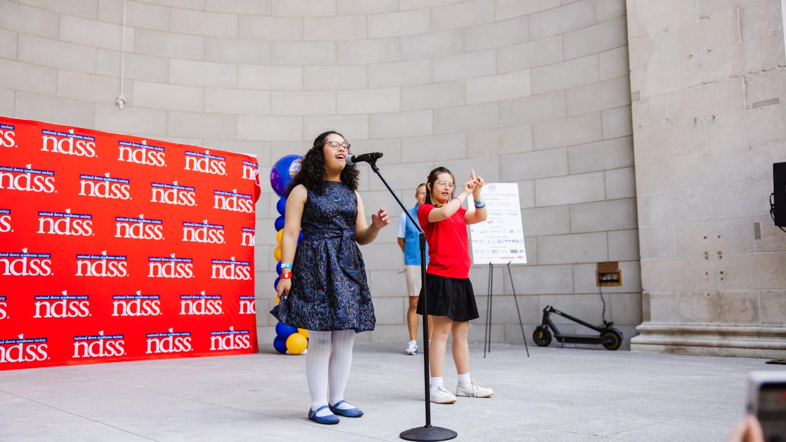 A young woman with Down syndrome, wearing glasses and a navy floral dress with white tights and blue flats, sings into a microphone on a stand. Behind her, a girl with Down syndrome in a red T-shirt and black skirt performs the song in American Sign Language, in the photo she is signing "stars". They stand in front of a red backdrop with a repeating NDSS logo, near a column of blue and gold balloons and a parked scooter on a stage.