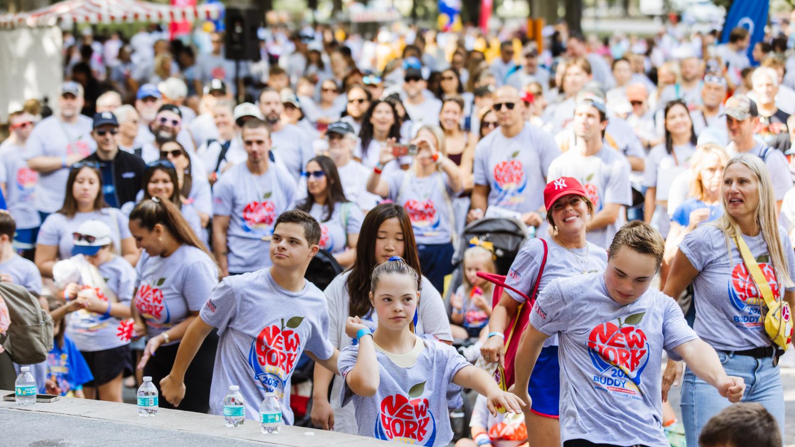A crowd of people wearing matching gray T-shirts printed with a red apple design and "New York City 2026 Buddy Walk" dance together outdoors. In the front row, a boy, a girl with Down syndrome, and another boy with Down syndrome dance with their arms extended.