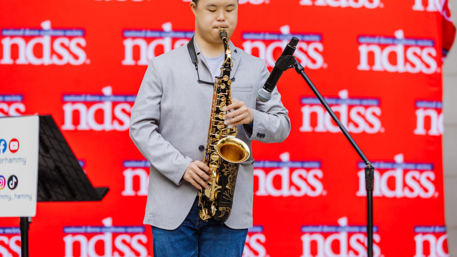 A young man with Down syndrome, wearing a gray blazer over a white T-shirt and jeans, plays a saxophone on an outdoor stage. He stands beside a microphone stand in front of a red backdrop with a repeating NDSS logo, with a sign displaying social media icons visible to his left.