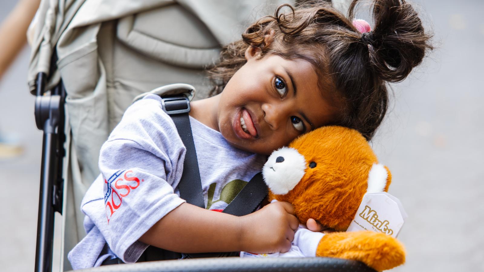 A young girl with curly hair styled in a bun rests her head on an orange-and-white stuffed bear while seated in a stroller. She wears a gray T-shirt with a partial NDSS logo printed on the sleeve and smiles.