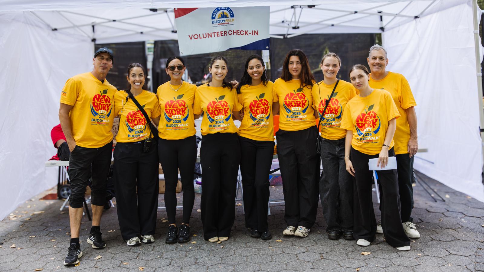 Eight adults stand together under a white event tent, all wearing matching yellow T-shirts printed with a red apple design and "New York City 2026 Buddy Walk, Educate | Advocate | Celebrate." Behind them hangs a sign reading "New York City Buddy Walk, Volunteer check-in."