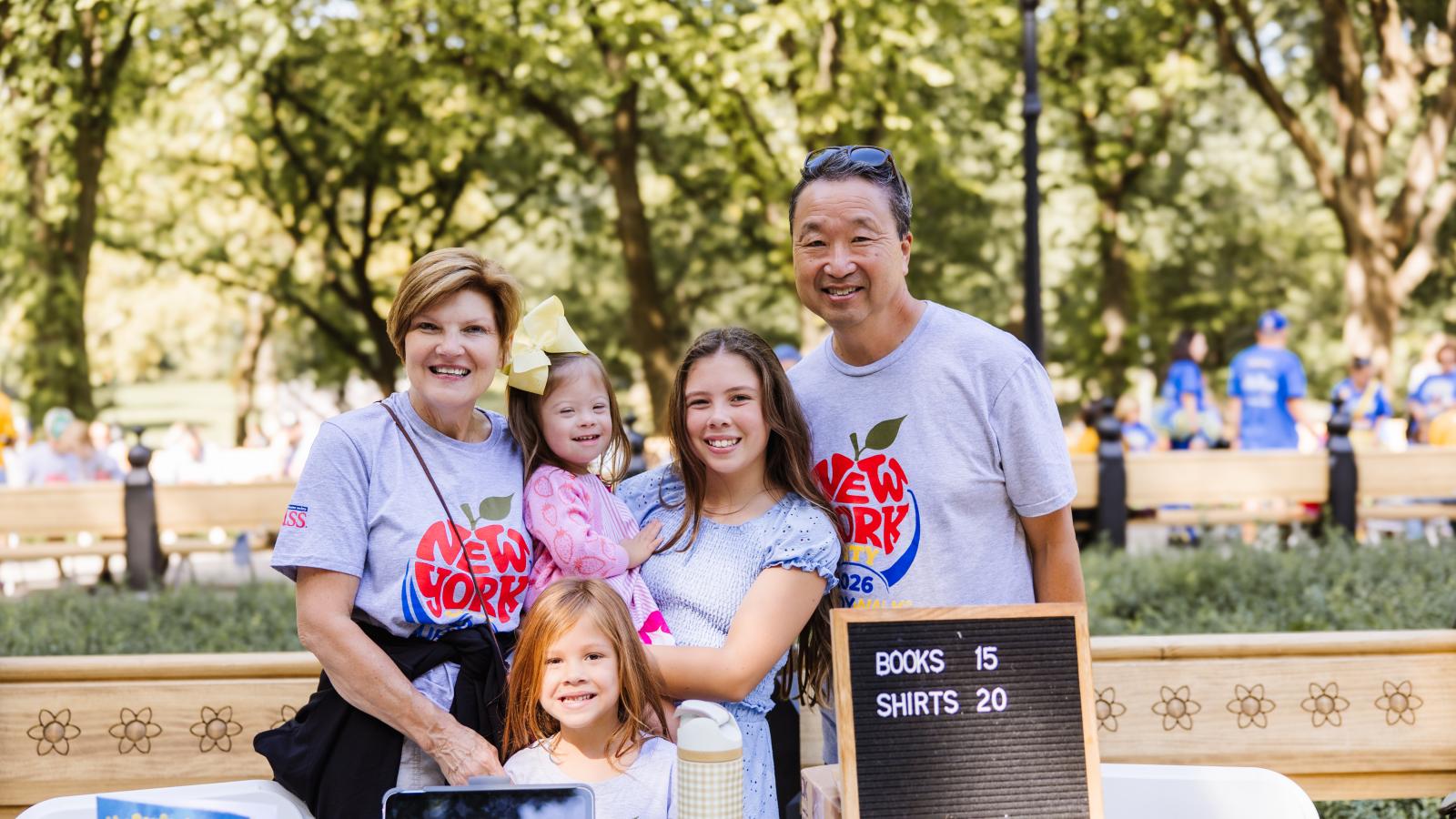 A woman with short hair, a man, and three girls (one with Down syndrome wearing a yellow bow) stand together outdoors behind a table, all in matching gray T-shirts printed with a red apple design and "New York City 2026 Buddy Walk." On the table are a tablet, a water bottle, a book titled "My Perfect Design," and a letter board reading "Books 15, Shirts 20."