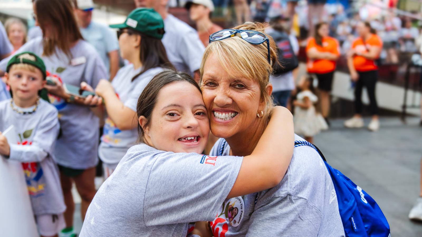 Close-up of a young woman with Down syndrome hugging an older woman at a crowded outdoor event. The young woman has dark hair and wears a gray T-shirt with a partial red, white and blue logo and a button reading "Layla's Zig Zaggers" with a photo. The woman has blonde hair, sunglasses pushed up on her head, gold hoop earrings, and a blue backpack slung over one shoulder. Both are smiling.