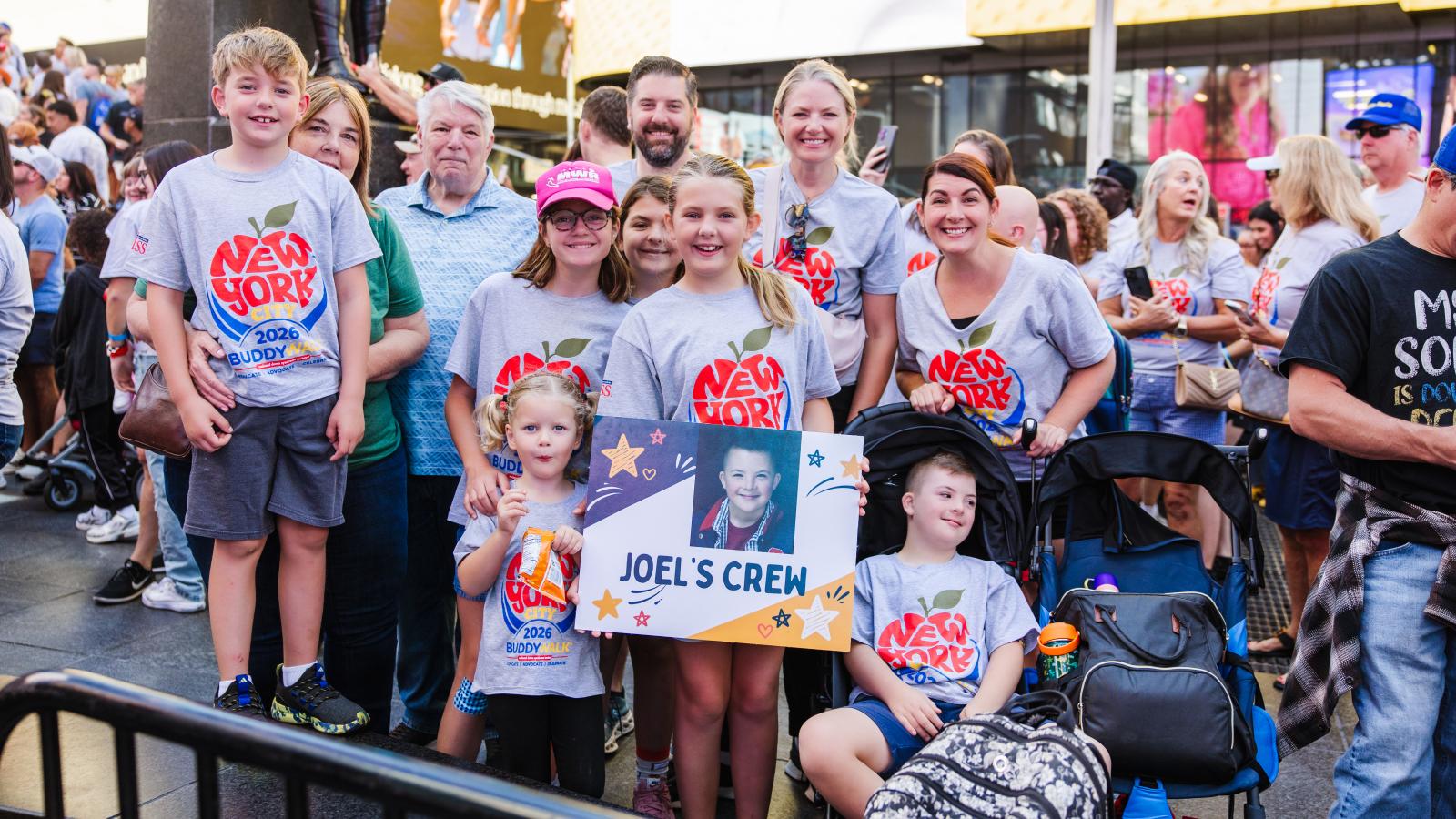 A group of about 10 people, including children and adults, pose together outdoors near a statue, most wearing gray T-shirts with a red, white and blue apple logo reading "New York City 2026 Buddy Walk, Educate Advocate Celebrate." A boy with Down syndrome sits in the front wearing the same shirt. Two young girls hold a sign reading "Joel's Crew" with a photo of a smiling boy in a red plaid shirt. Strollers and bags sit nearby.