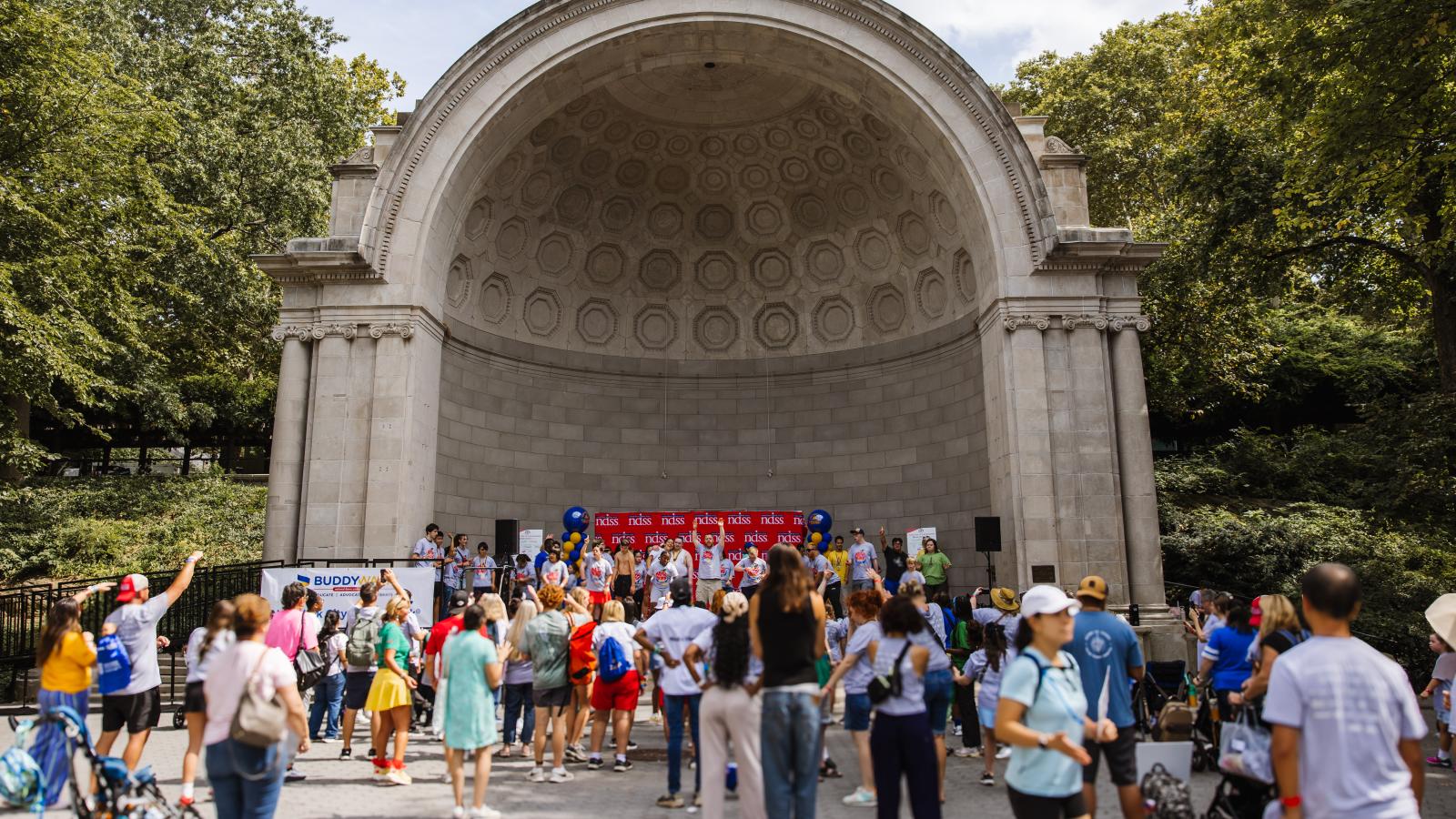 A crowd gathers outdoors in front of a large stone amphitheater with a domed, coffered ceiling. On a stage beneath the arch, a group in matching T-shirts stands in front of a red backdrop with a repeating NDSS logo, flanked by blue and gold balloon columns. A white banner in front reads "Buddy Walk" with additional text below it. People in the foreground, wearing various colored T-shirts, dance and raise their arms.