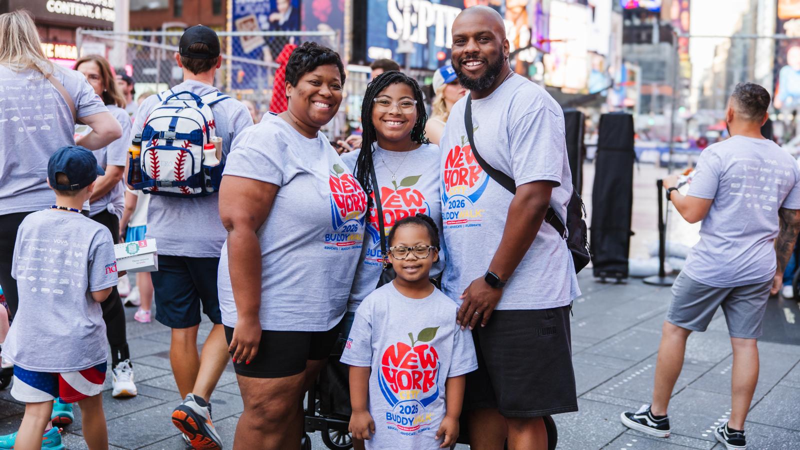 A woman, a teenage girl and a man stand together in Times Square, all wearing gray T-shirts with a red, white and blue apple logo reading "New York City 2026 Buddy Walk." In front of them, a girl with Down syndrome wearing glasses and the same T-shirt smiles at the camera. Illuminated billboards and a crowd fill the background in Times Square.