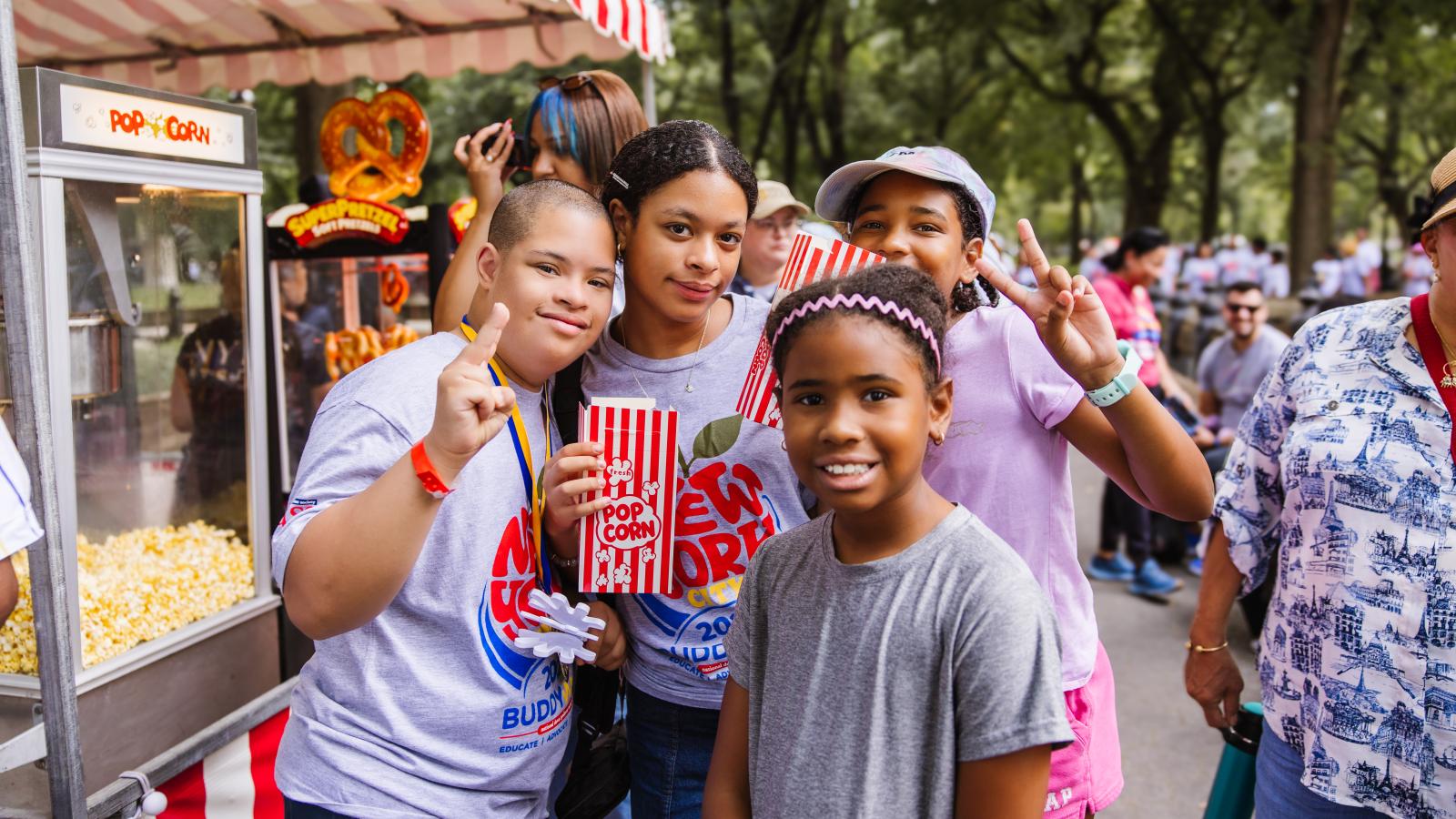 Four children stand together outside near a popcorn cart and a pretzel stand under a red-and-white striped awning. Two boys wear gray T-shirts printed with "New York City Buddy Walk, Educate | Advocate," one of them a boy with Down syndrome holding up a peace sign and a popcorn box. A girl in a light purple T-shirt and cap also holds up a peace sign, and a younger girl in a gray T-shirt with a pink headband smiles in front.